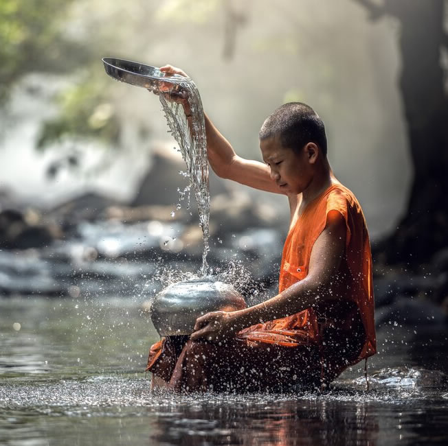 Junger buddhistischer Mönch in orangefarbener Robe sitzt in einem Fluss und gießt Wasser aus einer Metallschale in ein Gefäß, umgeben von friedlicher Natur.